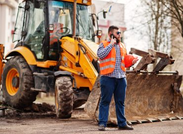 Beard worker man suit construction worker in safety orange helmet, sunglasses against tractor with mobile phone at hand.