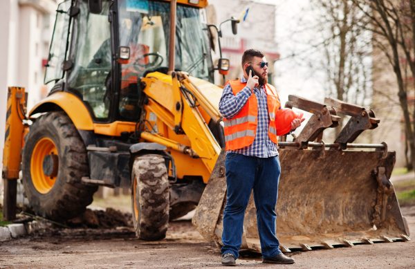 Beard worker man suit construction worker in safety orange helmet, sunglasses against tractor with mobile phone at hand.